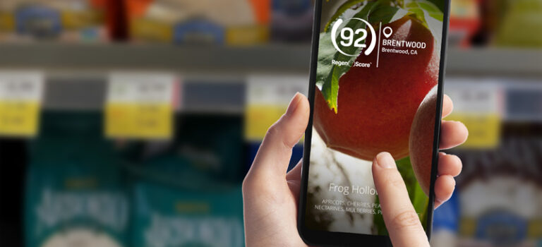 Hand holding smartphone with a blurred image of a supermarket aisle in background and finger swiping across screen that shows a closeup of a peach with RegenScore of 92 additional text to the side and below that says Frog Hollow, Brentwood CA