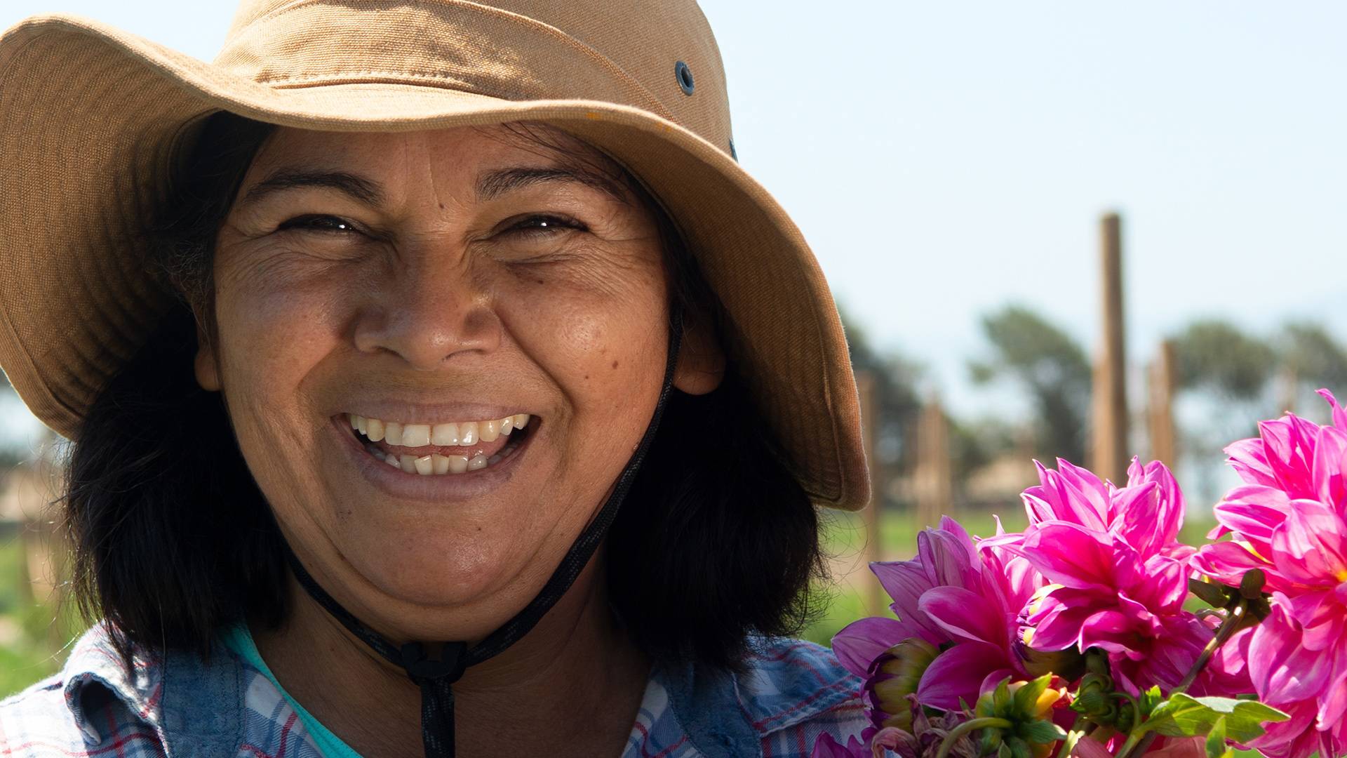 Closeup of smiling woman with hat in field  with posts in backgroundnext to fuschia colored flowers